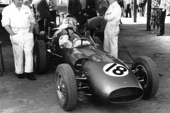Black-and-white photograph of the Aston Martin DBR5 in the pits in 1960, its driver at the wheel and mechanics in white overalls around it